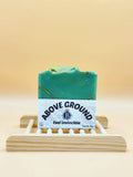 Green soap bar with 'Above Ground' label on a wooden stand against a beige background