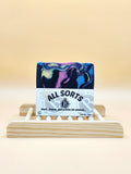 Colorful soap bar with 'All Sorts' packaging on a wooden stand against a beige background